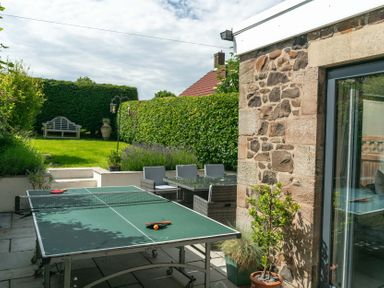 An outdoor patio with a ping pong table chairs and table a stone wall and a garden with a bench at Whinstones at 4 The Villas Embleton