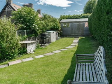 A garden with a stone path a bench a barbecue and a wooden shed at Whinstones at 4 The Villas in Embleton