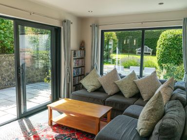 A living room with a gray sofa with patterned pillows a wooden coffee table and glass sliding doors overlooking a garden at Whinstones at 4 The Villas in Embleton
