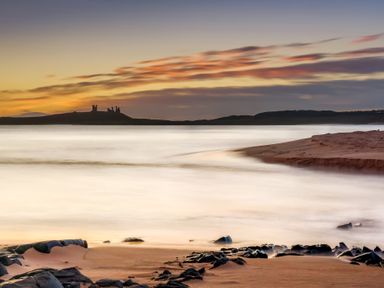 A sandy beach with rocks and water at sunset with castle ruins on a distant hill at Whinstones at 4 The Villas in Embleton