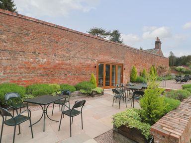 An outdoor patio area with tables and chairs next to a long brick wall with glass doors and surrounding plants at Lakeside Village Lodges & The Potting Shed in Capesthorne near Macclesfield