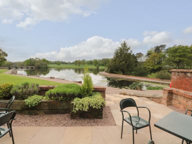 An outdoor patio with chairs and a table overlooking a pond and green landscape at Lakeside Village Lodges & The Potting Shed in Capesthorne near Macclesfield