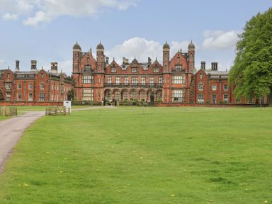 A large red brick mansion with multiple chimneys and towers in front of a green lawn with a driveway and trees at Lakeside Village Lodges & The Potting Shed in Capesthorne near Macclesfield