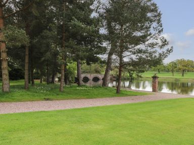 A grassy area with a pathway leading to a pond with trees and a stone bridge at Lakeside Village Lodges & The Potting Shed in Capesthorne near Macclesfield