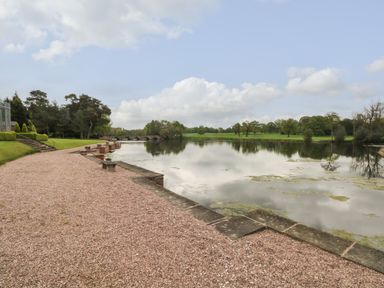A gravel path along a lake with stone benches and a stone bridge in the background at Lakeside Village Lodges & The Potting Shed Capesthorne near Macclesfield