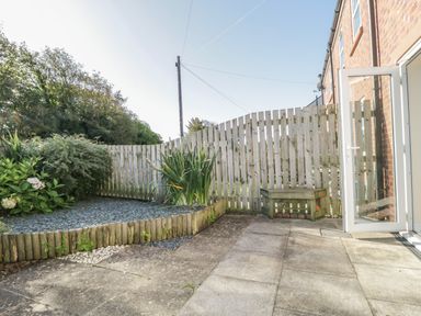 A patio with concrete paving next to a garden bed with plants and a wooden bench at Rowan Cottage in St Bees