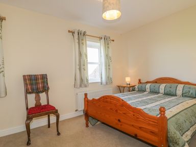 A bedroom with a wooden bed and green patterned bedspread a wooden chair with a plaid blanket and window with patterned curtains at Rowan Cottage in St Bees