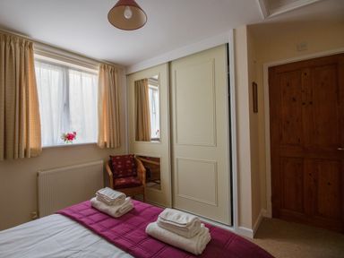 A bedroom with a bed with folded towels on a purple blanket a window with curtains a wooden door a wicker chair and a wardrobe with sliding doors with mirrors at Little Barn in Cucklington near Wincanton