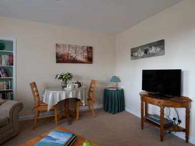 A living room with a small dining table with flowers and a tea set two wooden chairs a TV on a wooden stand a lamp on a round table and bookshelves at Little Barn in Cucklington near Wincanton