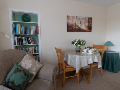 A living room with a sofa and cushions a small dining table with chairs and flowers and a bookshelf at Little Barn in Cucklington near Wincanton