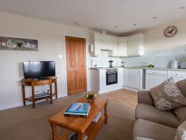A living area with a sofa and pillows, wooden coffee table with books and fruit bowl, a kitchen with white cabinets and appliances, and a wooden door at Little Barn Cucklington near Wincanton