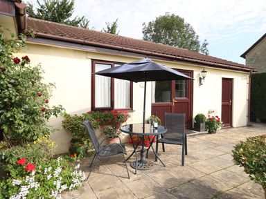 An outdoor patio with a round table umbrella and two chairs outside a cream building with brown doors and windows surrounded by plants and flowers at Little Barn Cucklington near Wincanton
