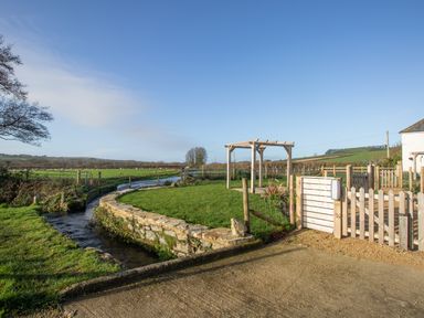 A garden with a wooden pergola a stone wall and a stream running beside a wooden gate at Willowbrook Cottage in Litton Cheney