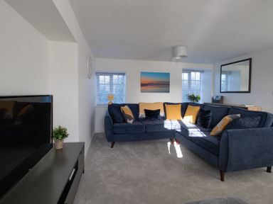A living room with a blue sectional sofa with yellow and blue cushions a television on a black stand and windows with blinds at Willowbrook Cottage in Litton Cheney