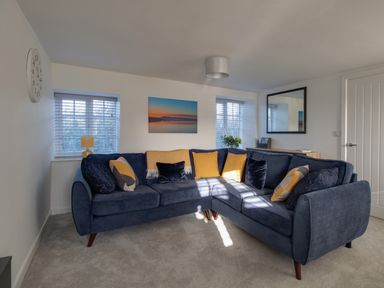 A living room with a blue sectional sofa with yellow and dark pillows two windows with blinds a wall clock a landscape picture a mirror and a wooden cabinet at Willowbrook Cottage in Litton Cheney