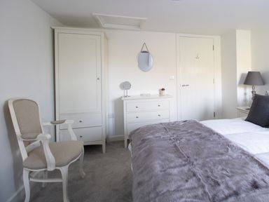A bedroom with a bed covered with a gray blanket a beige armchair a white wardrobe a chest of drawers with a small round mirror on top a door and a table lamp at Willowbrook Cottage in Litton Cheney