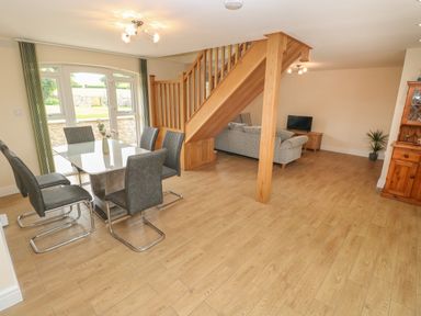 A dining area with a glass table and six gray chairs next to a staircase with a living room in the background at Woodpecker Barn in Hensol near Pontyclun