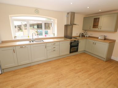 A kitchen with light wood flooring, grey cabinets, a stainless steel oven, stovetop, and sink under a large window at Woodpecker Barn in Hensol near Pontyclun