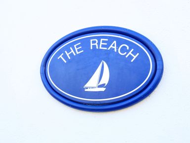 A blue oval sign with a white sailboat and the words The Reach on a white wall at The Reach in Hallsands