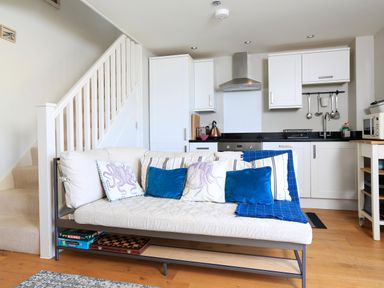 A small open plan kitchen with white cabinets and a white sofa with blue and striped cushions in front at The Reach in Hallsands