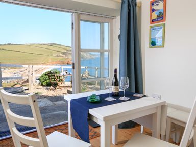 A dining area with a white table set for two and a view of a seaside patio with chairs and hills at The Reach in Hallsands