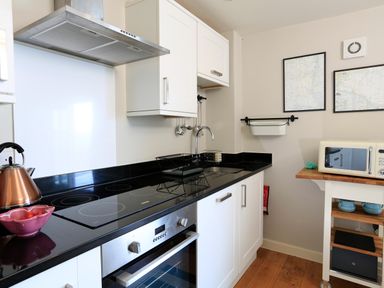 A kitchen with white cabinets a black countertop an electric stove a stainless steel oven a copper kettle a microwave on a wooden cart and framed maps on the wall at The Reach in Hallsands