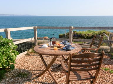 A wooden table set with breakfast items and two wooden chairs on a gravel patio overlooking the sea at The Reach in Hallsands