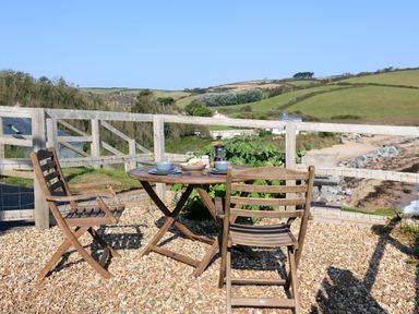 An outdoor wooden table and chairs set on gravel with cups and plates near a wooden fence overlooking hills and a beach at The Reach in Hallsands