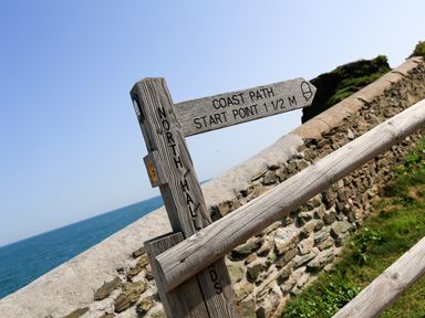 A wooden signpost near a stone wall by the sea indicating coast path start point at The Reach in Hallsands