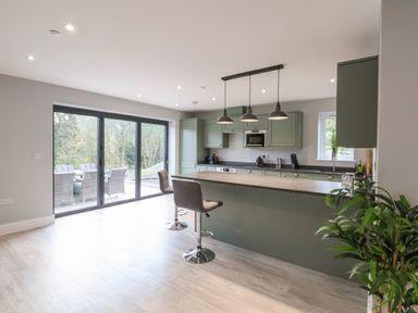 A kitchen with green cabinets an island with two bar stools pendant lights and sliding glass doors leading to an outdoor seating area at Fron Haul - Robin Goch Llanfwrog near Ruthin