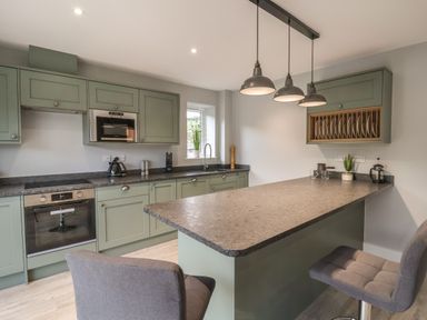 A kitchen with green cabinets a countertop with two chairs and hanging lights at Fron Haul - Robin Goch in Llanfwrog near Ruthin
