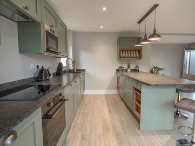 A kitchen with green cabinets a gray countertop a stovetop an oven a microwave hanging lights and bar stools at Fron Haul - Robin Goch in Llanfwrog near Ruthin