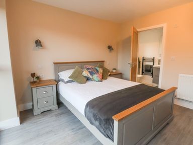 A bedroom with a bed with pillows and a blanket two bedside tables with plants wall lights an open door leading to a bathroom at Fron Haul - Robin Goch in Llanfwrog near Ruthin