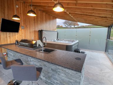 An outdoor covered area with a bar counter a sink two stools a barbecue a hot tub and a wall-mounted television at Fron Haul - Robin Goch Llanfwrog near Ruthin