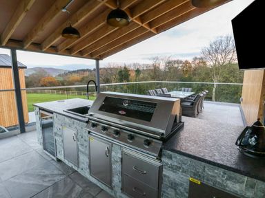 An outdoor kitchen with a built-in grill sink and countertop with dining table and chairs on a covered patio at Fron Haul - Robin Goch Llanfwrog near Ruthin