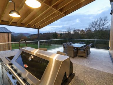 An outdoor patio with a stainless steel barbecue grill a dining table with chairs and a glass railing overlooking trees and grass at Fron Haul - Robin Goch in Llanfwrog near Ruthin