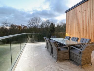 An outdoor terrace with a glass railing a dining table and wicker chairs at Fron Haul - Robin Goch Llanfwrog near Ruthin