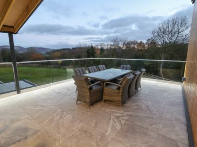 An outdoor patio with a glass dining table and eight wicker chairs surrounded by glass railing at Fron Haul - Robin Goch in Llanfwrog near Ruthin