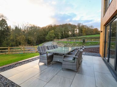 An outdoor patio with a glass table and eight cushioned chairs near a wooden building at Fron Haul - Robin Goch in Llanfwrog near Ruthin