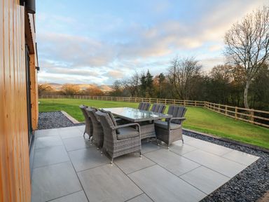 An outdoor patio with a glass-top table and eight cushioned chairs overlooking a grassy yard with a wooden fence and trees at Fron Haul - Robin Goch Llanfwrog near Ruthin
