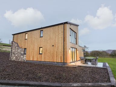 A wooden house with large windows and outdoor seating area in a rural setting at Fron Haul - Robin Goch in Llanfwrog near Ruthin