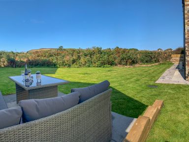 An outdoor patio with a wicker sofa and glass table on a concrete surface overlooking a grassy lawn and hedge at Beacon House in St Agnes