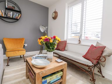 A living room with a yellow chair a wooden coffee table with flowers and a magazine a bench with red pillows and a bookshelf on a grey wall at Beacon House in St Agnes