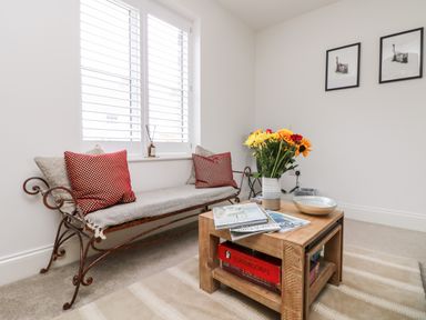 A sitting area with a metal framed bench with cushions a wooden coffee table with flowers and magazines on it and two framed pictures on the wall at Beacon House in St Agnes