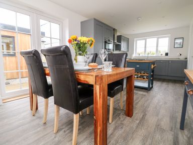 A dining area with a wooden table and black chairs next to a kitchen with grey cabinets and a kitchen island at Beacon House in St Agnes