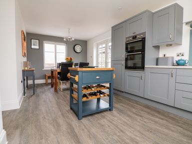 A kitchen with gray cabinets a blue kitchen island with wine bottles and a dining area with a wooden table and black chairs at Beacon House in St Agnes