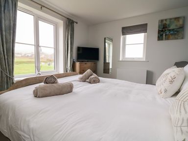 A bedroom with a bed and rolled towels on the bed near windows with curtains and a television on a dresser at Beacon House in St Agnes