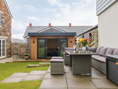 An outdoor patio with wicker furniture including a table and cushioned seating a small wooden shed and green lawn at Beacon House in St Agnes