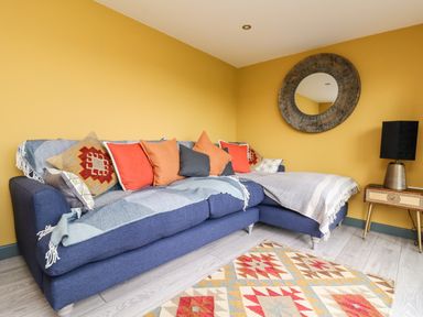 A living room with a blue sofa and colorful cushions a patterned rug and a round mirror on a yellow wall at Beacon House in St Agnes