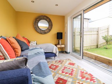 A living room with a blue sofa and orange cushions a round wall mirror a floor lamp a patterned rug and glass sliding doors opening to a garden at Beacon House in St Agnes